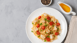 Imagem de um prato com uma porção de espaguete com bacalhau, ao lado uma pequena jarra de azeite e um potinho de pimenta do reino.